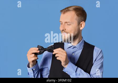 Jeune homme souriant barbu portant une chemise bleue et un gilet noir, tenant un nœud papillon noir, isolé sur un fond bleu Banque D'Images