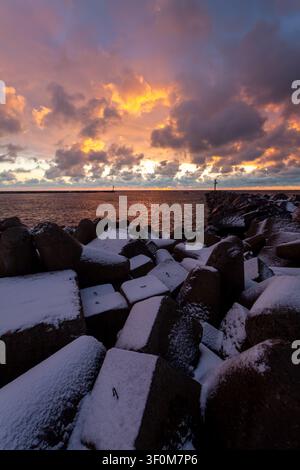 Rocher enneigés au bord d'une mer calme sous un ciel rougeoyant au coucher du soleil avec des nuages dramatiques, créant une scène côtière hivernale paisible. Banque D'Images