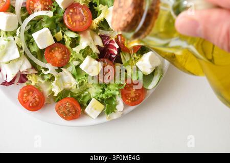 Salade mixte fraîche avec des tomates cerises, des cubes de feta, des oignons, des feuilles vertes et de l'huile d'olive versée sur les ingrédients dans une composition en gros plan lumineux Banque D'Images