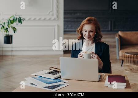 Femme aux cheveux rouges boit du café tout en travaillant sur ordinateur portable avec des écouteurs dans le bureau à domicile moderne Banque D'Images