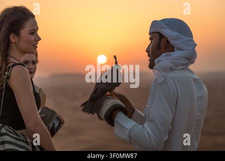 Un fauconnier émirati vêtu d'une kandoura blanche traditionnelle et d'un keffiyeh présente un faucon encapuchonné sur un gant à une touriste féminine dans un désert. Banque D'Images