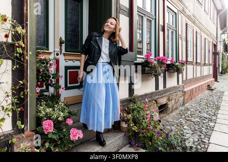 Modèle femme souriante debout près d'une porte sur une rue pavée, portant une veste en cuir, une jupe bleue et des bottes. Fleurs roses, façade en bois et Banque D'Images