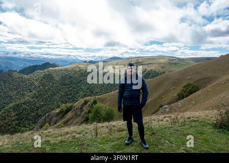 Un randonneur vêtu d'une doudoune bleu marine et d'un bonnet se tient sur une crête de prairie alpine dans le Caucase. Banque D'Images