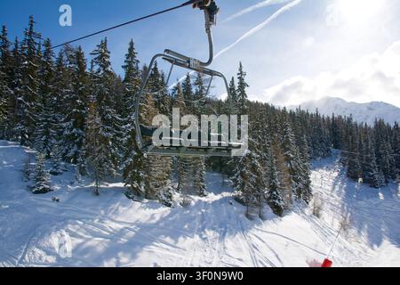 Un téléphérique à quatre places vide glisse au-dessus d'une piste de ski damée bordée par des épicéas et des sapins chargés de neige dans une station de ski alpine. Banque D'Images