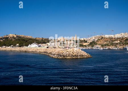Vue d'arrivée du port de Mgarr à Gozo, avec un phare de brise-lames, une ferry, une marina, le village de Ghajnsielem, l'église Notre-Dame de Loreto et t Banque D'Images