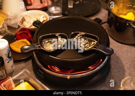 Vue rapprochée d'un grill à raclette avec de petites poêles individuelles, des restes de fromage fondu, des pommes de terre et de la vaisselle lors d'un dîner privé dans un endroit non identifié a Banque D'Images