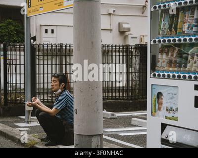 Homme accroupi à côté d'un poteau électrique en fumant une cigarette à côté d'un distributeur de boissons dans une rue de Shibuya, Tokyo, Japon Banque D'Images