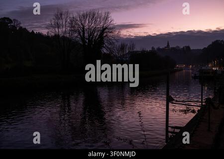 Vue de la rivière Rance au crépuscule avec des silhouettes d'arbres sombres, des quais, un ciel coucher de soleil violet-rose et un clocher d'église lointain près du port de Dinan à Dinan Banque D'Images