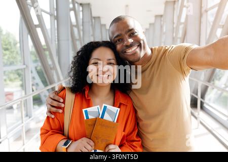 Couple noir souriant prenant un selfie dans un couloir lumineux de l'aéroport tout en tenant leurs passeports Banque D'Images
