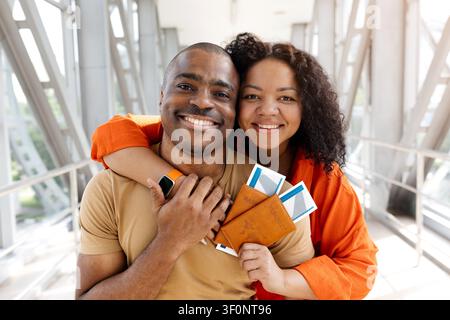 Portrait d'un couple noir souriant qui s'embrasse dans un couloir d'aéroport tout en tenant leurs passeports Banque D'Images