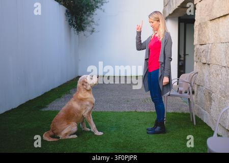 Femme instruisant un chien Goldendoodle en dressage à l'extérieur d'une maison moderne Banque D'Images