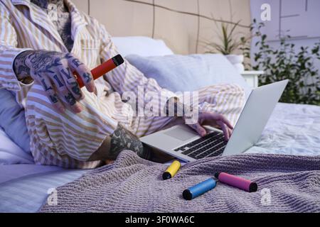 Jeune homme beau en pyjama avec différentes cigarettes électroniques jetables et ordinateur portable sur le lit à la maison Banque D'Images