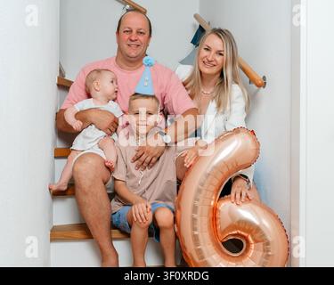 Une famille de quatre personnes célèbre joyeusement sur un escalier en bois, orné de ballons et de chapeaux de fête, capturé en gros plan, avec un angle légèrement surélevé Banque D'Images
