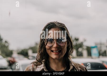 Femme lunettes de pluie jeune femme aux cheveux mouillés souriant debout sous la pluie par une journée nuageuse Banque D'Images