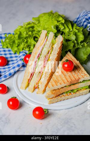 Une assiette contient un sandwich avec des couches de jambon, de fromage et de laitue. Des mini-tomates fraîches et de la laitue verte entourent le plat. Ceci est servi sur un damier Banque D'Images
