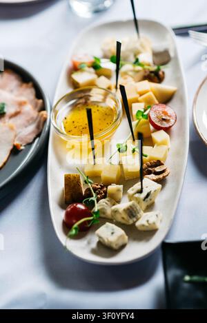 Planche à fromage élégante mettant en vedette une variété de fromages à pâte dure et bleus accompagnés de miel, de raisins rouges, de noix, de micro-pousses fraîches et de pics à cocktail sur un plat en céramique blanche. Apéritif raffiné magnifiquement présenté sur une table de restaurant. Banque D'Images