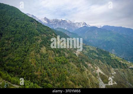 Une vue aérienne de pentes montagneuses couvertes d'une forêt dense avec des sommets enneigés au loin dans l'Himachal Pradesh. Banque D'Images