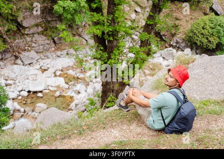 Un jeune homme avec un sac à dos est assis sur une pente herbeuse surplombant un ruisseau de montagne rocheux dans l'Himalaya. Banque D'Images