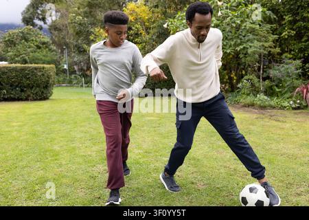 Père afro-américain et fils adolescent pratiquant le football dans le jardin en vêtements de sport avec un ballon de football Banque D'Images