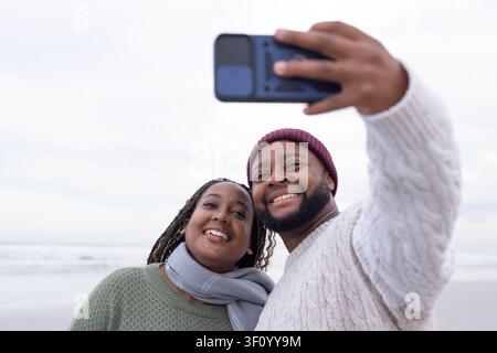 Couple africain souriant et posant pour un selfie sur une plage de sable, tenant un smartphone et des pulls en tricot Banque D'Images