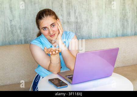 Jeune femme avec ordinateur portable offrant une main tout en travaillant en ligne à domicile et souriant à l'objectif Banque D'Images