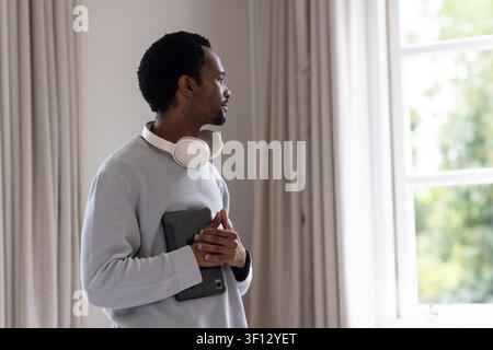 Homme debout à gauche près d'une fenêtre à multiples vitres regardant au-dehors, tenant une tablette et portant des écouteurs Banque D'Images