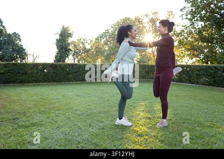 Diverses amies féminines s'étirant et se tenant en équilibre sur une jambe sur une pelouse en herbe avec des vêtements de sport, au soleil Banque D'Images