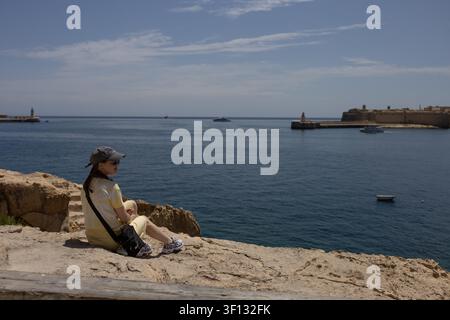 Femme assise sur une falaise rocheuse et regardant la baie du Grand Harbour à La Valette, Malte, le 19 juin 2026 Banque D'Images