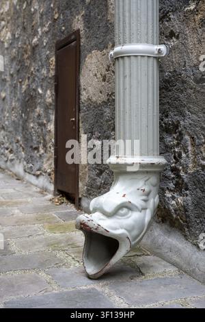 Vue détaillée d'un bec de gouttière antique en forme de gargouille décorative. Le tuyau métallique décoré dirige l'eau vers le sol pavé Banque D'Images