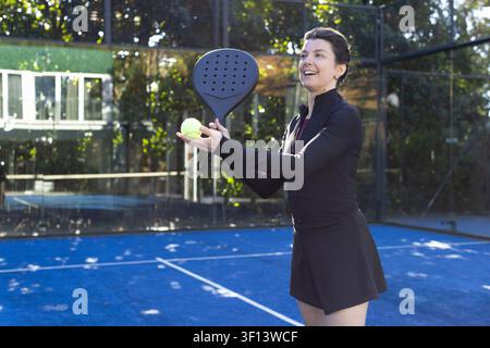 Femme debout sur un court de padel bleu portant une tenue noire tenant une raquette et une balle jaune, espace de copie Banque D'Images
