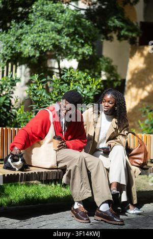 Couple afro-américain positif, homme et femme, se détendant ensemble assis sur un banc en caressant un chat errant. Banque D'Images