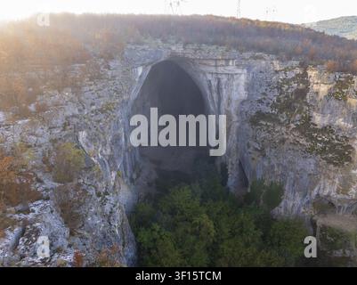 Vue aérienne de la grotte Prohodna présentant son immense arche de calcaire, des arbres verdoyants à l'entrée et des formations rocheuses accidentées sous un matin doux l Banque D'Images