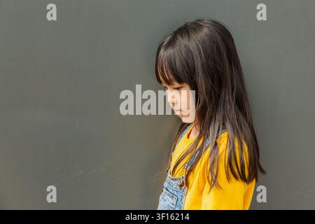 Jeune fille asiatique d'âge scolaire pensive de profil, debout seule et regardant vers le bas contre un mur de béton gris simple Banque D'Images