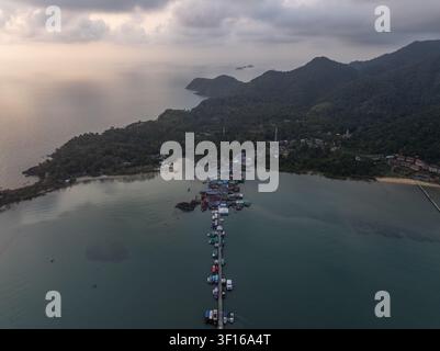Vue aérienne du quai de Bang Bao s'étendant dans la mer avec un village de pêcheurs traditionnel, des bateaux amarrés et des collines verdoyantes sous un ciel doux au coucher du soleil à Banque D'Images