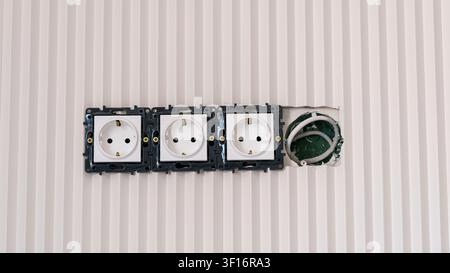 Trois prises électriques sont visibles sur un mur, dont une prise partiellement installée, révélant le câblage et une surface murale texturée dans un environnement domestique Banque D'Images