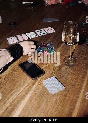 Table en bois lors d'une partie de poker avec des cartes à jouer, des jetons colorés, un verre de vin blanc et un smartphone. Concept de luxe pour un passe-temps. vertical Banque D'Images