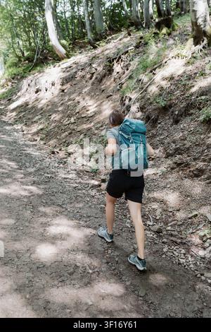 Vue arrière d'une randonneuse portant un grand sac à dos bleu, marchant sur un sentier forestier escarpé par une journée ensoleillée Banque D'Images