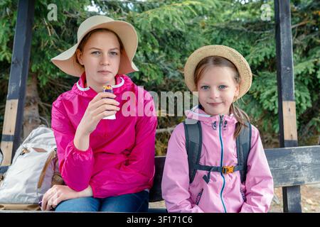 Deux filles prenant une pause snack pendant une randonnée, assises sur un banc en bois dans la forêt portant des chapeaux de soleil et des vestes roses, l'une mangeant une barre énergétique, con Banque D'Images