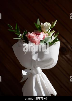 Un petit bouquet élégant avec des roses roses et blanches, des eustoma et de l'eucalyptus enveloppés dans du papier texturé blanc avec un ruban Banque D'Images