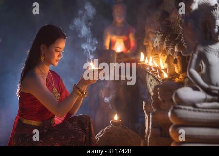 Vue de côté d'une jeune Thaïlandaise en tenue traditionnelle accroupie devant une rangée de statues de Bouddha dans un temple avec une bougie Banque D'Images