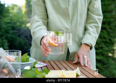 Grossissement d'une femme debout près d'une table de jardin tenant un verre d'eau glacé avec des pamplemousses fraîches et des feuilles de menthe Banque D'Images