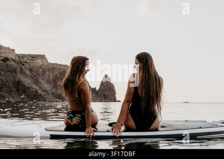 Women Paddleboard Beach Sunset - deux femmes s'assoient sur une planche de paddleboard dans l'océan au coucher du soleil, se regardant l'une l'autre. Banque D'Images