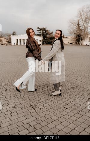 Amies femmes marchant ensemble à travers une place pavée de la ville tout en portant des manteaux d'automne élégants pendant des vacances de voyage Banque D'Images