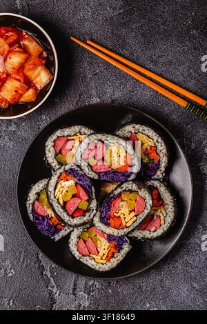 Une assiette noire remplie de kimbap (ou gimbap) aux garnitures colorées. Les ingrédients comprennent des saucisses, des cornichons, des œufs et des légumes. Banque D'Images
