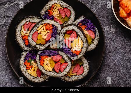 Une assiette noire remplie de kimbap (ou gimbap) aux garnitures colorées. Les ingrédients comprennent des saucisses, des cornichons, des œufs et des légumes. Banque D'Images