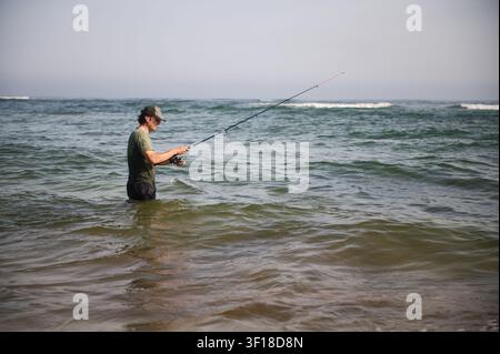 Un homme se tient jusqu'à la taille dans la mer, lançant une canne à pêche dans des vagues douces, profitant d'une journée tranquille de pêche en surf et de détente en plein air le long de la côte Banque D'Images