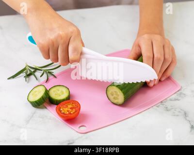 Mains d'enfant coupant un concombre avec un couteau en plastique sécurisé sur une planche à découper rose avec tomate et romarin en gros plan Banque D'Images