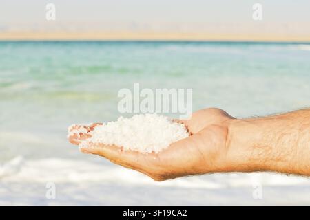 Granules de sel de la Mer Morte dans les mains sur la plage de Jordanie, gros plan de minéraux étincelants devant une eau bleue. Sel riche en minéraux récolté depuis le point le plus bas sur Terre Banque D'Images
