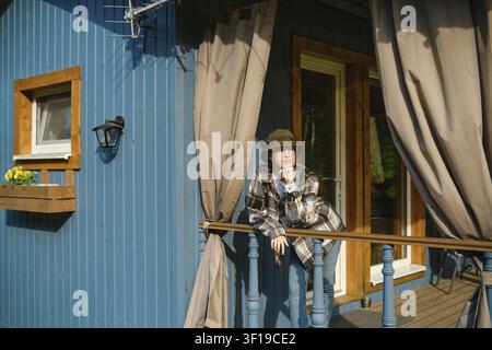 Jeune femme pensive appuyée sur la rambarde d'une cabane en bois bleue, portant un chapeau et une chemise à carreaux, regardant pensivement au loin. Bright sunlight shines on he Banque D'Images