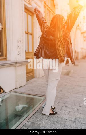 Femme célébration rue. Femme joyeuse aux bras levés debout sur une rue pavée au coucher du soleil, baignée par la lumière dorée de la ville Banque D'Images